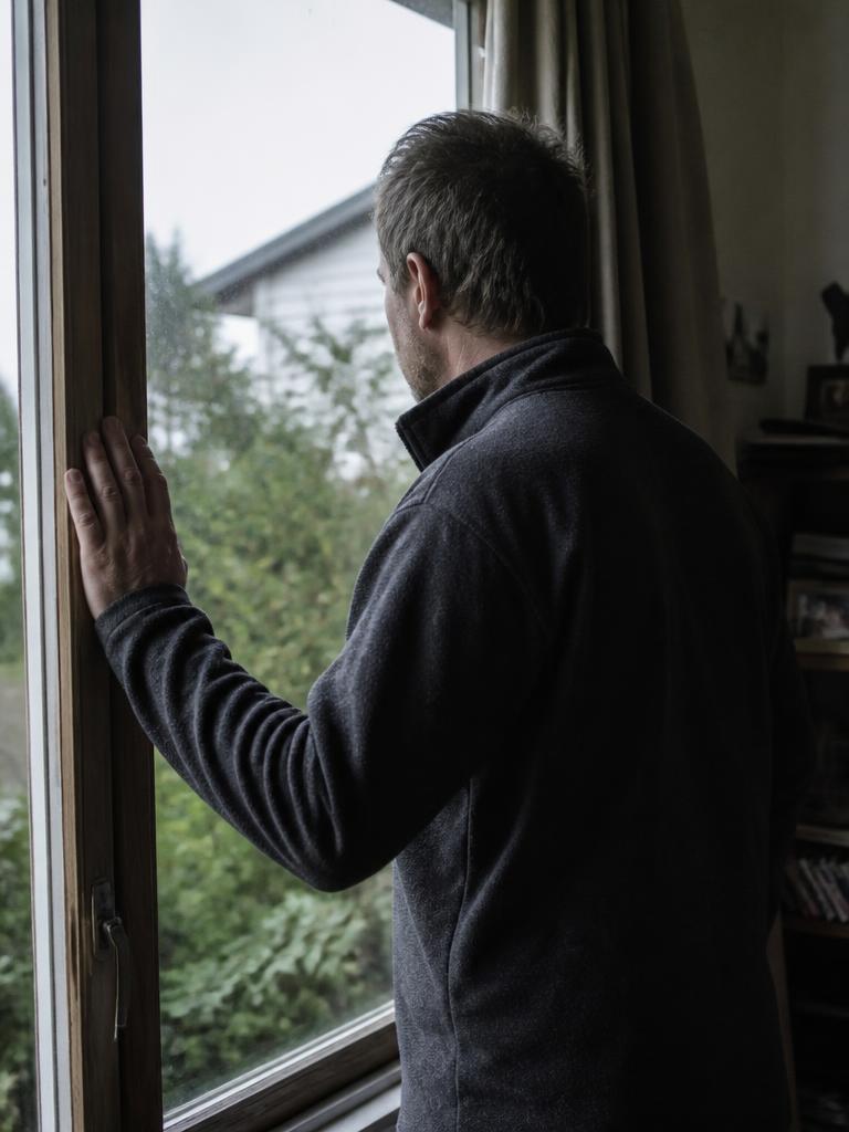 A man in his late forties standing at a window in a New Zealand home, looking outside.